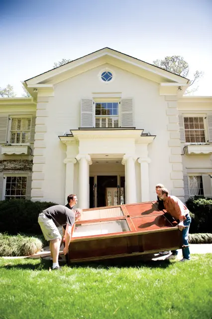 Two men carrying a large wooden display cabinet with glass doors in front of a white brick house with columns and shutters.