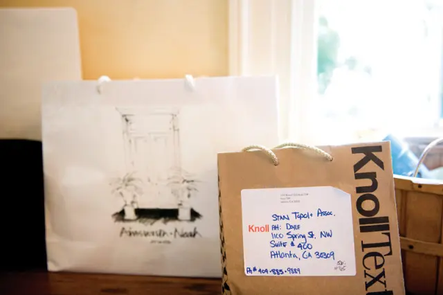 Two shopping bags on a wooden surface. The white bag on the left has a sketch of a doorway and plants. The brown bag on the right has the KnollText logo and a shipping label with an address and phone number.