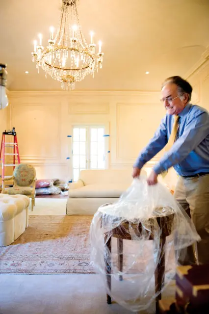 Man in blue shirt and yellow tie covering a wooden table with plastic wrap in a room with a chandelier, sofa, armchair, and ladder.