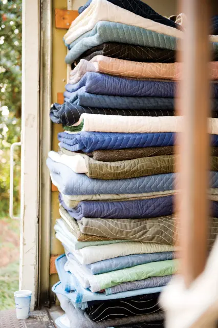 Stack of quilted moving blankets in various shades of blue, green, gray, and cream, with white stitching in diamond and chevron patterns.