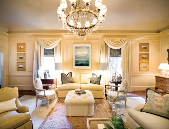 Living room with cream walls, a large crystal chandelier, a cream sofa, two patterned armchairs, and a tufted ottoman on an oriental rug.