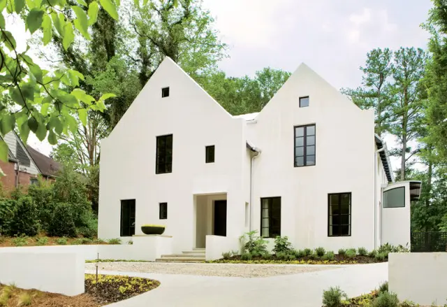 Modern white stucco house with asymmetrical gabled roofs, black-framed windows, and a gravel driveway surrounded by lush green trees.