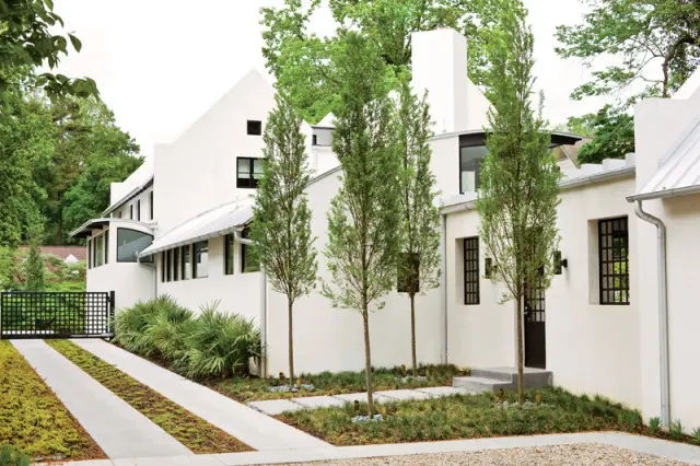 Modern white stucco house with metal roof, tall trees, and a driveway leading to a black metal gate.
