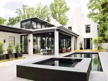 Modern white stucco house with black trim and large windows, featuring a covered patio with outdoor dining and seating, and a rectangular swimming pool and hot tub in the courtyard.