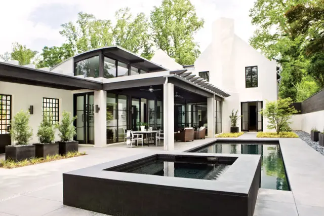 Modern white stucco house with black trim and large windows, featuring a covered patio with outdoor dining and seating, and a rectangular swimming pool and hot tub in the courtyard.