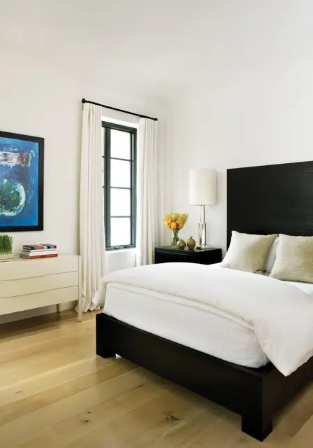 Bedroom with a dark wood platform bed, white bedding, two textured pillows, a black nightstand with a lamp and vase of yellow flowers, and a white dresser with a framed abstract painting.