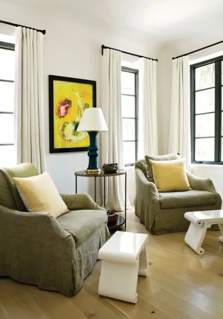Living room with two slipcovered armchairs in olive green, yellow throw pillows, abstract yellow artwork, and white accent tables on a light wood floor.