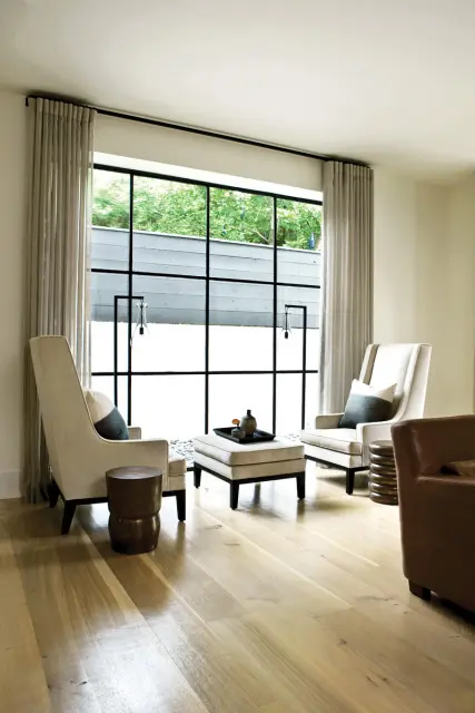Living room with two cream upholstered armchairs facing a large window with a black grid. A cream ottoman sits between the chairs, holding a tray with decorative objects.