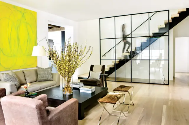 Modern living room with a large yellow abstract painting, taupe sofa, dark wood coffee table, and a glass-enclosed staircase with a person ascending.
