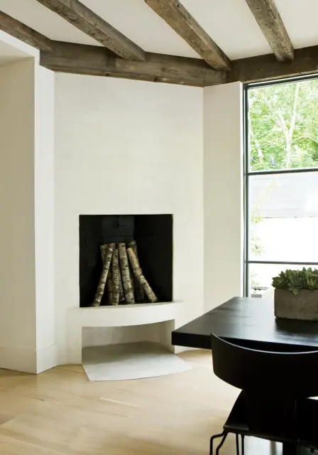 Modern living room with white plaster fireplace, exposed wooden beams, black dining table, and large window overlooking greenery