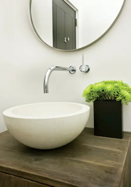 Powder room with a stone vessel sink on a rustic wood vanity, chrome wall-mounted faucet, and a black planter filled with green mums. A round mirror reflects a dark gray door.