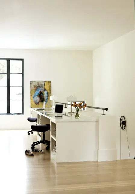 Modern white built-in desk with laptops and two black rolling chairs, featuring a large window and abstract art on the wall.