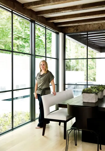 Woman standing by a large window in a dining room with a dark wood table, white banquette seating, and concrete planters with succulents.