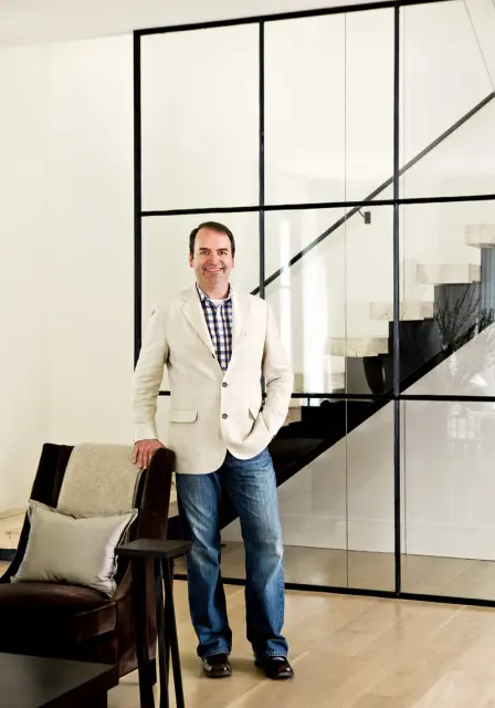 Man in a light-colored blazer and jeans standing in a modern interior with a glass-enclosed staircase behind him.