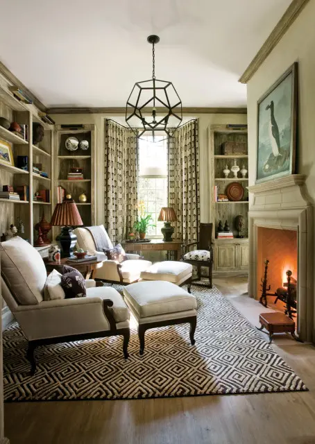 Living room with two upholstered armchairs and ottomans on a geometric rug, flanked by bookshelves and a fireplace with a painting above.