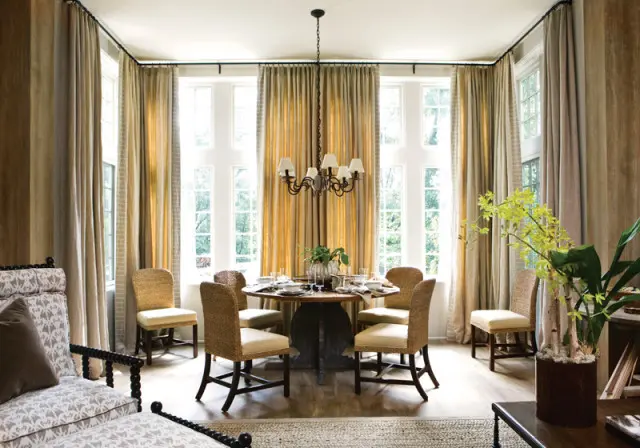 Dining room with round wooden table set for six, surrounded by woven chairs with cream cushions. Tall windows are draped in gold and beige curtains.