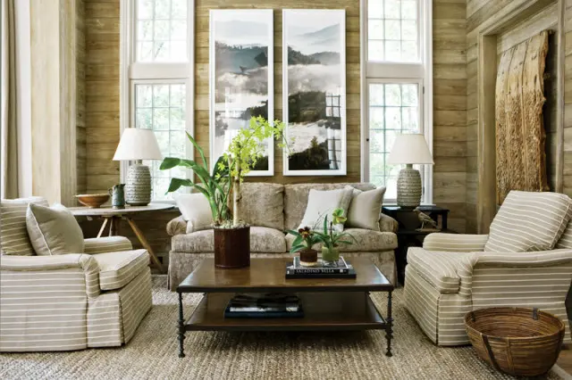 Living room with wood-paneled walls, two striped armchairs, a neutral sofa, a dark wood coffee table, and two framed landscape artworks above the sofa.