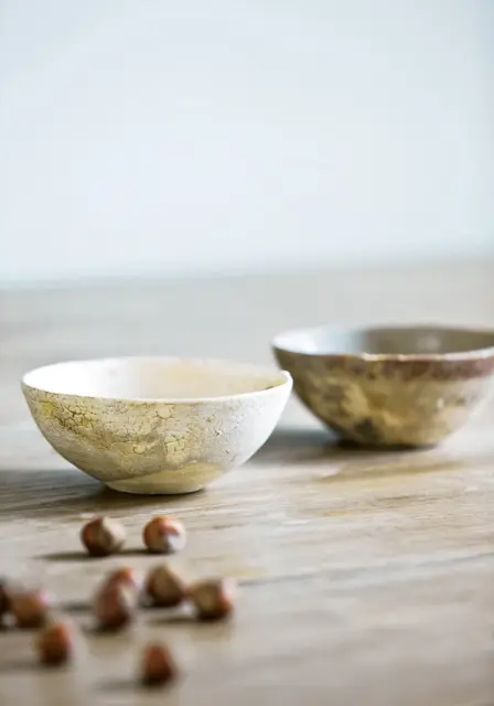 Two rustic ceramic bowls with textured, crackled finishes sit on a wooden surface, with hazelnuts scattered in the foreground.