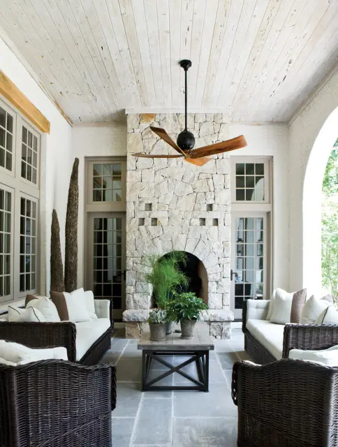 Covered patio with stone fireplace, white wood-planked ceiling, dark wicker furniture, and a wooden ceiling fan.