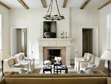 Living room with cream walls, exposed wooden beams, a stone fireplace with a circular chandelier above, and upholstered armchairs arranged around coffee tables.