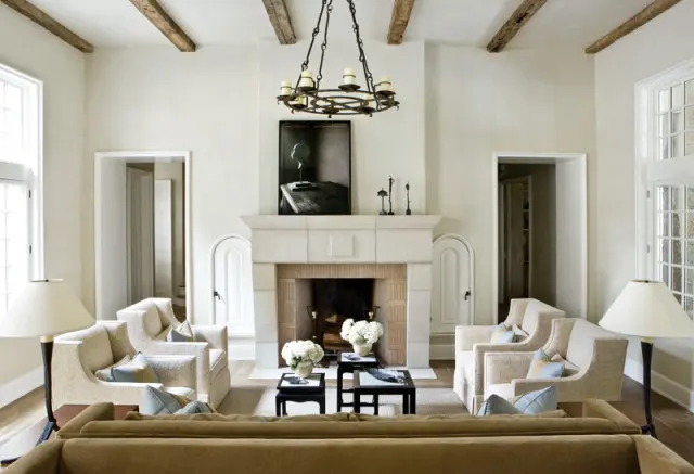 Living room with cream walls, exposed wooden beams, a stone fireplace with a circular chandelier above, and upholstered armchairs arranged around coffee tables.