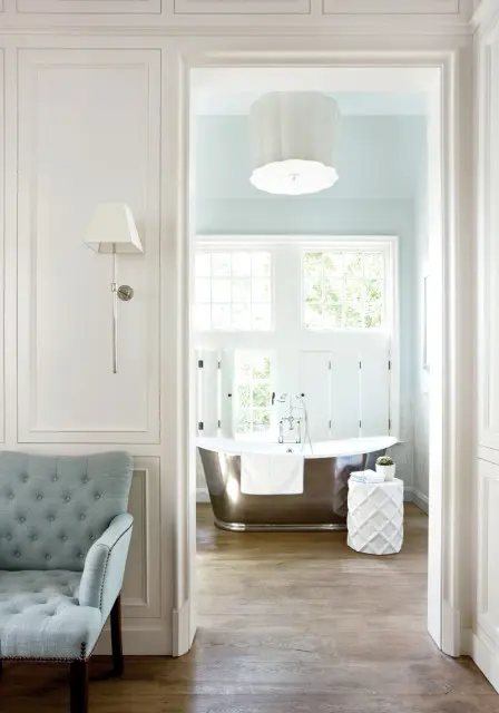 View through an archway into a light blue bathroom with a freestanding metallic tub, white shutters, and a large decorative ceiling light.