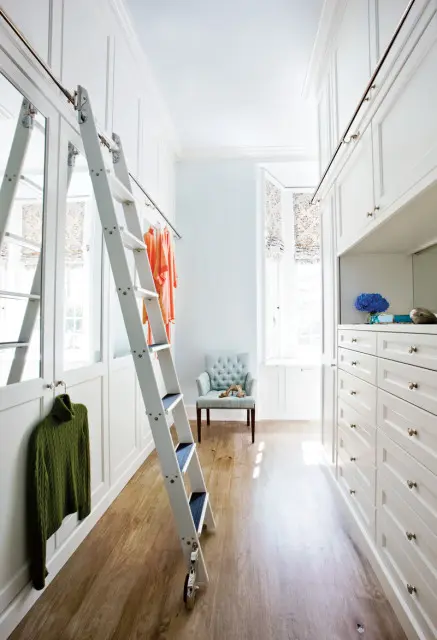 White walk-in closet with floor-to-ceiling custom cabinetry, a rolling ladder, tufted armchair, and hardwood floors.
