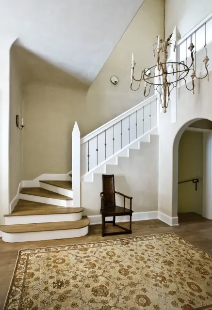 Foyer with a curved staircase featuring white spindles, a dark wood armchair, a large floral rug, and an ornate chandelier.