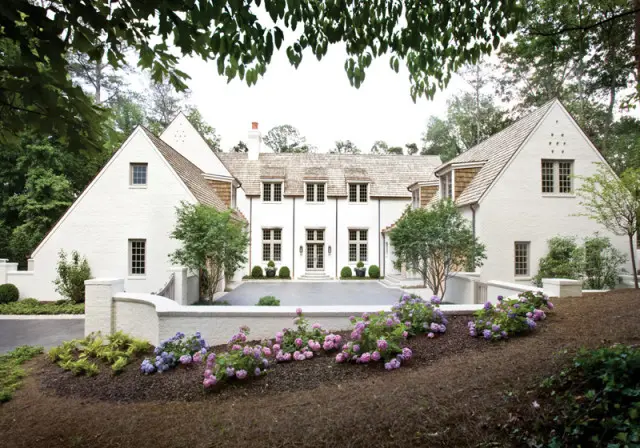 White stucco house with multiple gabled roofs, cedar shake accents, and numerous multi-paned windows, set amidst lush greenery and flowering bushes.