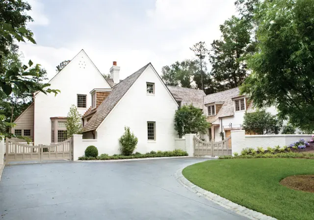 White brick house with wood shake roof, multiple gables, and light-colored wooden gates leading to a driveway and manicured lawn.
