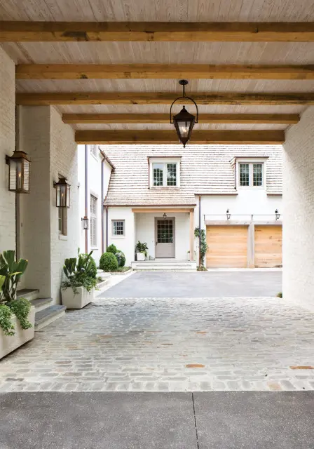 White brick house with a light gray front door, wooden garage doors, and a shingled roof, viewed from a covered entryway with exposed wooden beams and lanterns.