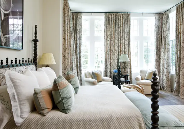 Bedroom with a dark wood four-poster bed, white bedding, patterned curtains, and two armchairs by the windows.
