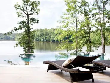 Infinity pool overlooking a lake surrounded by trees, with two dark wicker lounge chairs in the foreground.
