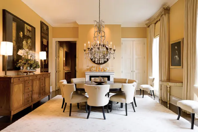 Dining room with a round dark wood table surrounded by upholstered chairs, a crystal chandelier, a fireplace, and a large wooden buffet with lamps and floral arrangement.