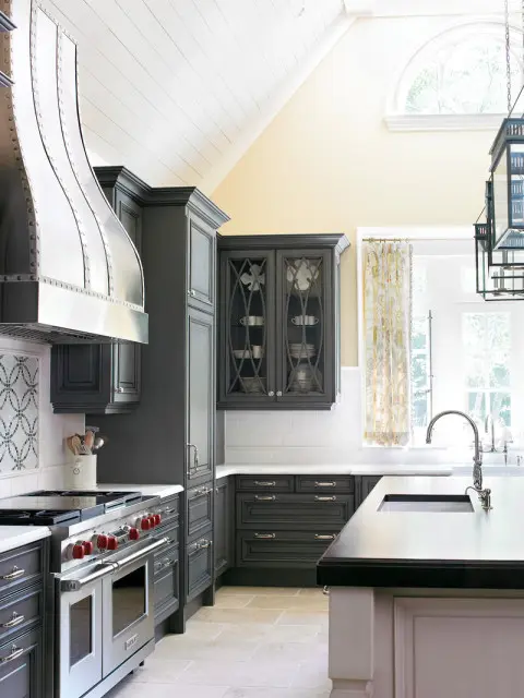 Dark gray kitchen cabinets with glass-front uppers, stainless steel range with red knobs, and a large metal range hood.