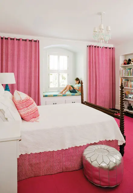 Girl reading on window seat in a bedroom with pink curtains, a white bed with a patterned skirt, and a silver pouf.