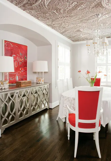 Dining room with a paisley patterned ceiling, mirrored buffet, abstract red art, and white chairs with red cushions around a round table.