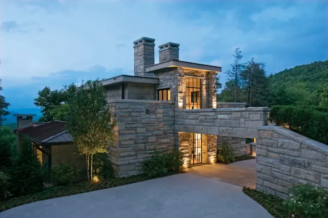 Modern stone house with multiple chimneys and large windows, set against a backdrop of lush green mountains at dusk.