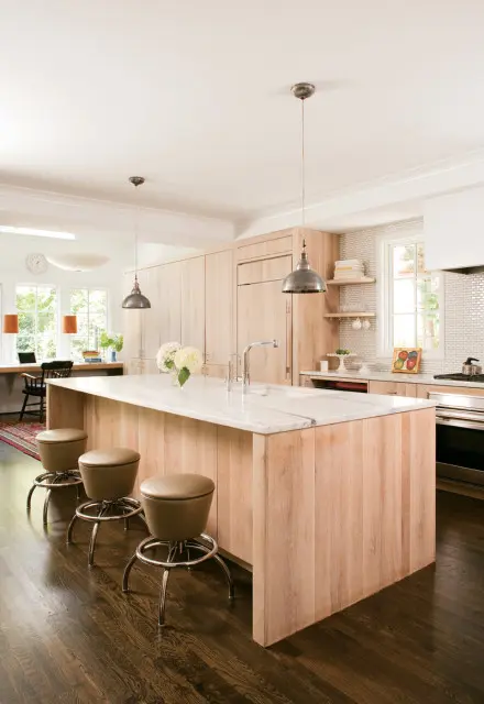 Light wood kitchen with a large marble island, stainless steel faucet, and three brown leather bar stools.