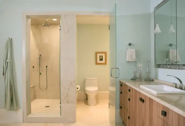 Light blue bathroom with marble shower stall, white toilet, and wood vanity with a white sink. A light green robe hangs on the wall.