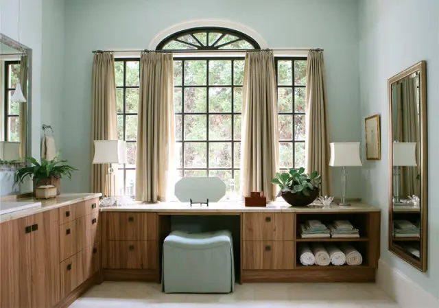 Light blue bathroom with a long wooden vanity featuring marble countertops, built-in drawers, and open shelving with rolled towels. A large arched window with grid panes overlooks greenery.