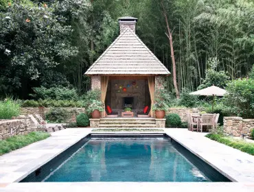 Rectangular swimming pool with stone patio leading to a covered outdoor living area with fireplace and seating, surrounded by lush bamboo and stone walls.