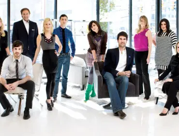 A group of nine professionals, men and women, posing in a modern office space with large windows.