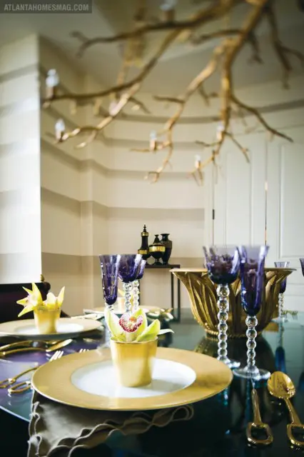 Dining table set with gold chargers, white plates, purple stemmed glasses, and gold vases with orchids. A gold branch-like chandelier hangs above.