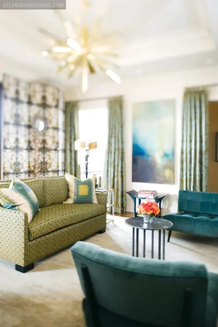 Living room with a green patterned sofa, teal upholstered chairs, and a starburst chandelier. Blue and yellow abstract art hangs on the wall.