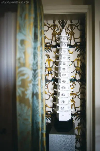White pagoda sculpture displayed against patterned wallpaper featuring figures in gold and teal. A patterned curtain hangs to the left.