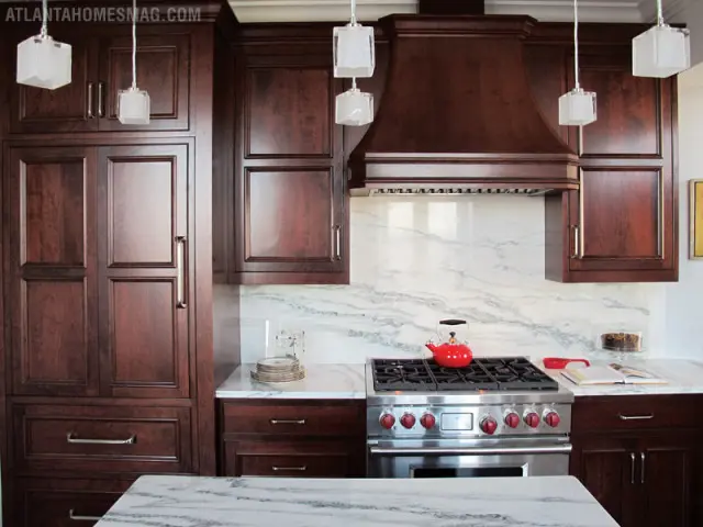 Dark cherry wood kitchen cabinets with marble countertops and backsplash, stainless steel range, and glass pendant lights.