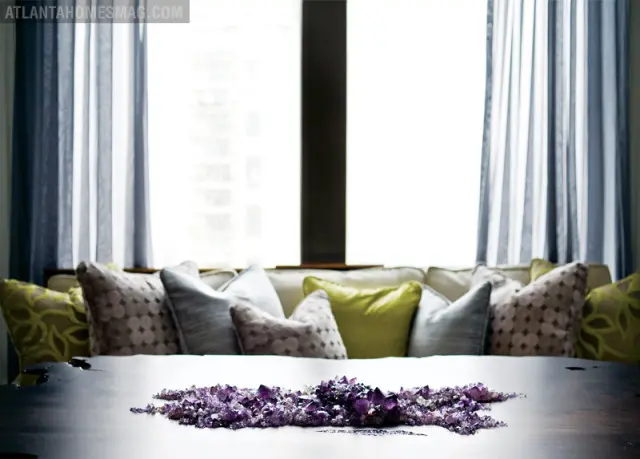 Dark wood table with amethyst crystal cluster centerpiece in front of a window with blue curtains and a sofa with patterned pillows.