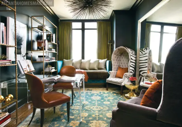 Dark teal study with a clear acrylic desk, brown leather chairs, gold shelving, a tufted high-back chair with zebra print, and a large ornate mirror.