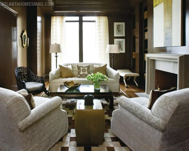 Living room with dark brown walls, white sofa, ornate black armchair, and two light-colored armchairs. Features a fireplace, built-in bookshelves, and a patterned rug.